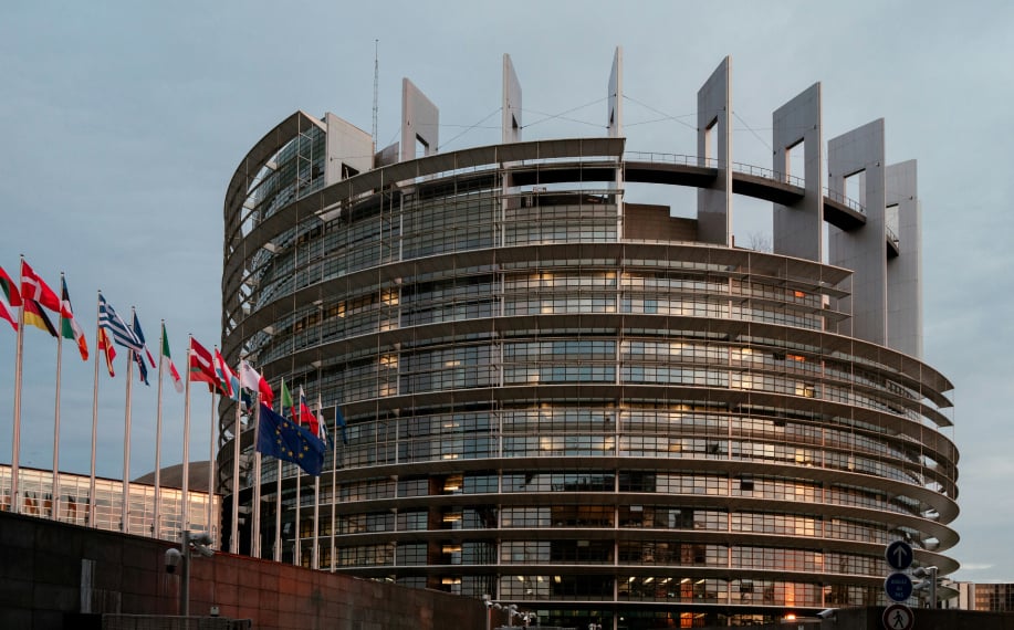 Euroopan Parlamenttirakennus kuvattuna vaaleansinisen taivaan alla. Liput liehuvat vierellä.