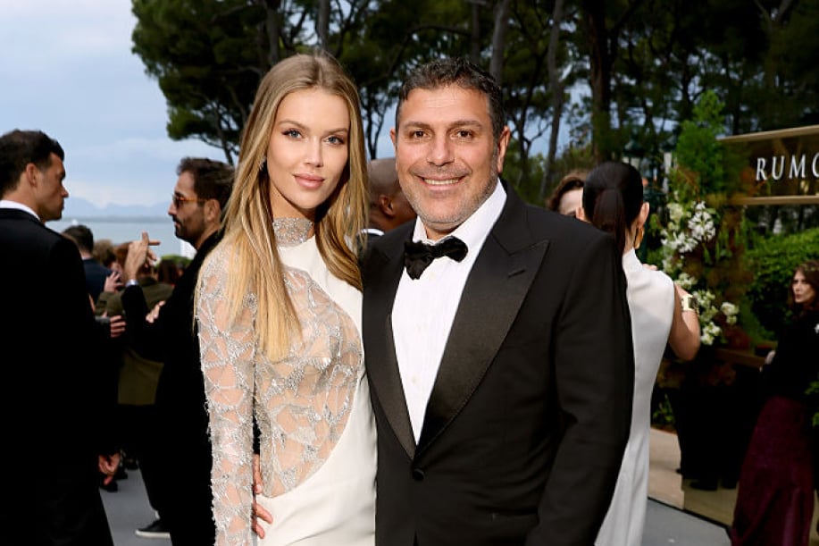 Juhlavaan iltapukutapahtumaan pukeutunut mies ja nainen poseeraavat yhdessä kameralle ulkoilmassa. Naisella on vaalea, yksityiskohtaisesti koristeltu iltapuku ja miehellä musta smokki. Taustalla näkyy muita vieraita, puita sekä merimaisema.