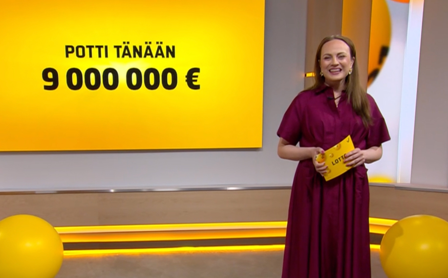 Televisiolähetyksen kuvakaappaus Lotto-arvonnasta, jossa juontaja seisoo Lotto-studiossa ja pitää arvontakuorta kädessään. Taustalla lukee “Potti tänään 9 000 000 €”, mikä kertoo kierroksen pääpotin suuruuden. Kuva liittyy lotto tuloksiin tänään, lotto arvontaan ja siihen, kuinka suuri potti on jaossa ennen lotto voitonjakoa.