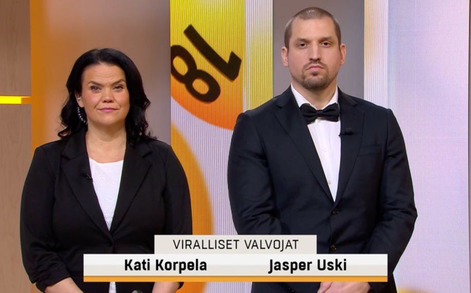 Kuvassa Lotto-arvonnan viralliset valvojat Kati Korpela ja Jasper Uski seisovat studiossa suorassa kuvakulmassa. He valvovat, että Lotto-arvonta toteutetaan sääntöjen mukaisesti ja että lotto tulokset sekä oikeat lottonumerot vahvistetaan luotettavasti ennen julkaisua. Taustalla näkyy Veikkauksen Lotto-lähetyksen tunnistettava keltainen ja valkoinen studiografiikka.