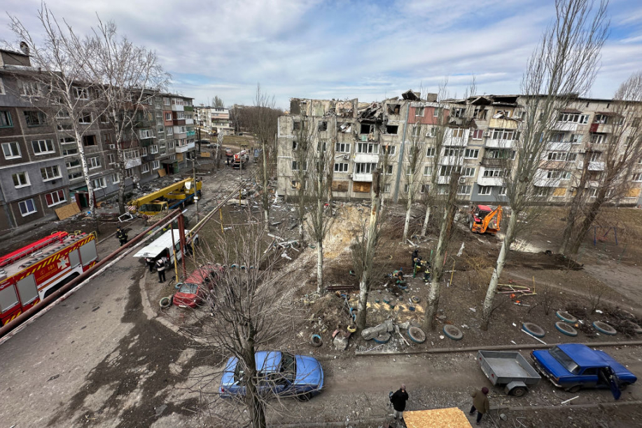 Asuntojen raunioita, autoja ja pelastushenkilökuntaa. Sininen hieman pilvinen taivas ja lehdettömoä puita. Karut pihat mitkä näyttävät osittain hiiltyneen.