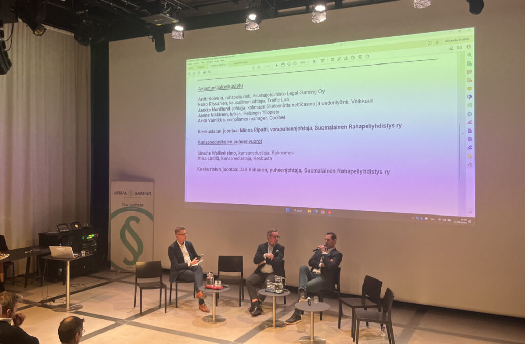 Kolme miestä lavalla keskuskirjasto Oodissa. Projektorin heijastus taustalla listaa puhujat tapahtumassa ketkä ovat Antti Koivula Asianajotoimisto Legal Gaming Oy, Esko Rissanen Traffic Lab, Jarkko Nordlund Veikkaus, Janne Nikkinen Helsingin Yliopistosta ja Antti Vainikka Coolbetiltä. Kansanedustajapuheenvuorot keskustan Mika Lintilällä ja kokoomuksen Sinuhe Wallinheimolla. Lisäksi mainitaan kskustelun juontaja Jari Vähänen Suomalaisesta Rahapeliyhdistyksestä.