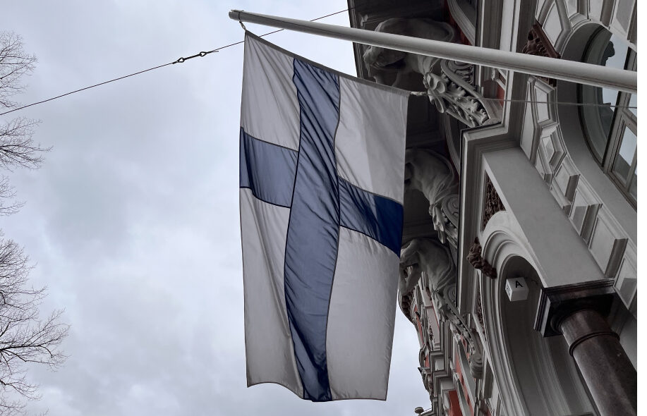 Suomen lippu tummanpuhuvalla taivastaustalla koreaa helsinkiläistä Jugend-rakennusta vasten.